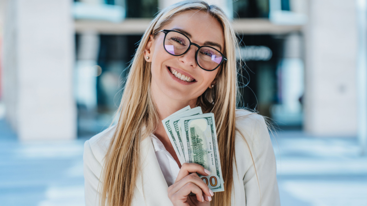Excited blonde caucasian woman in glasses, white suite holding US dollar banknotes smiling wide, satisfied by profit against blurry buildings.