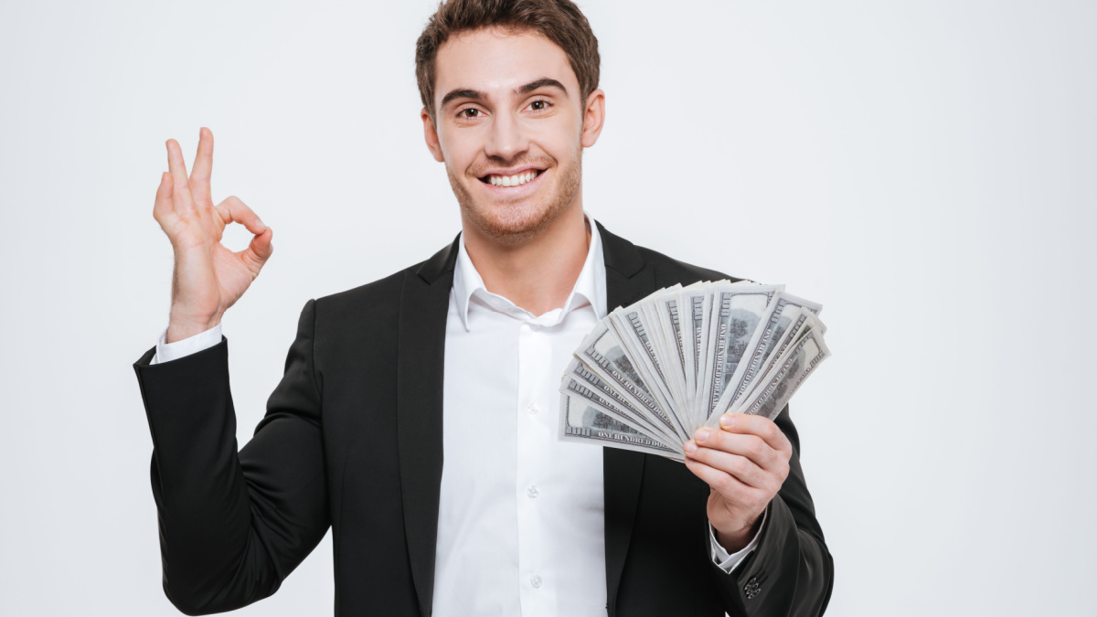 Photo of young cheerful businessman standing at studio holding money in hands make okay gesture. Isolated over white background.